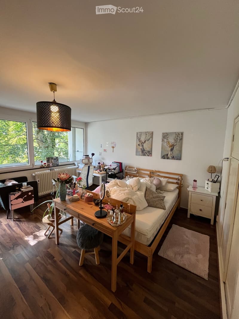 Cozy bedroom with a wooden bed, desk, and nightstand. Features a large window with a view of greenery, a hanging lamp, and decorative artwork on the walls.