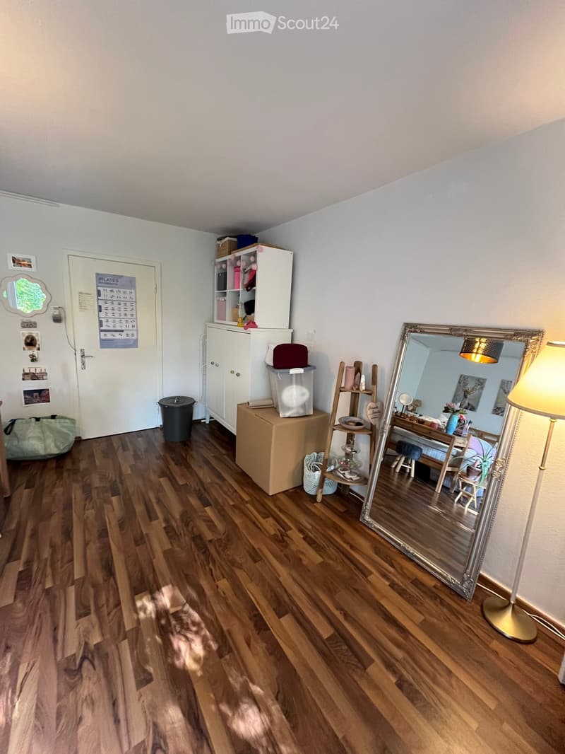 The room features wooden flooring, a large ornate mirror, a cabinet, a small table with a lamp, and several stools. There is also a door with a calendar and some pictures on the wall.