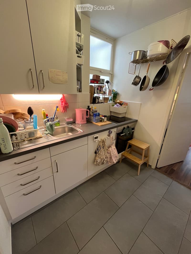 Modern kitchen with white cabinets, stainless steel sink, oven, and various kitchen utensils. The space includes a small wooden stool and hanging pots.