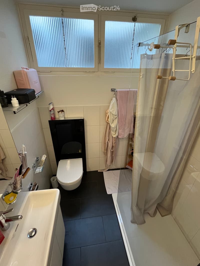 Modern bathroom with a white sink, chrome faucet, toilet, and a glass-enclosed shower with a curtain. Towels and a bathrobe are hanging on a rack, and various toiletries are placed on the sink counter.