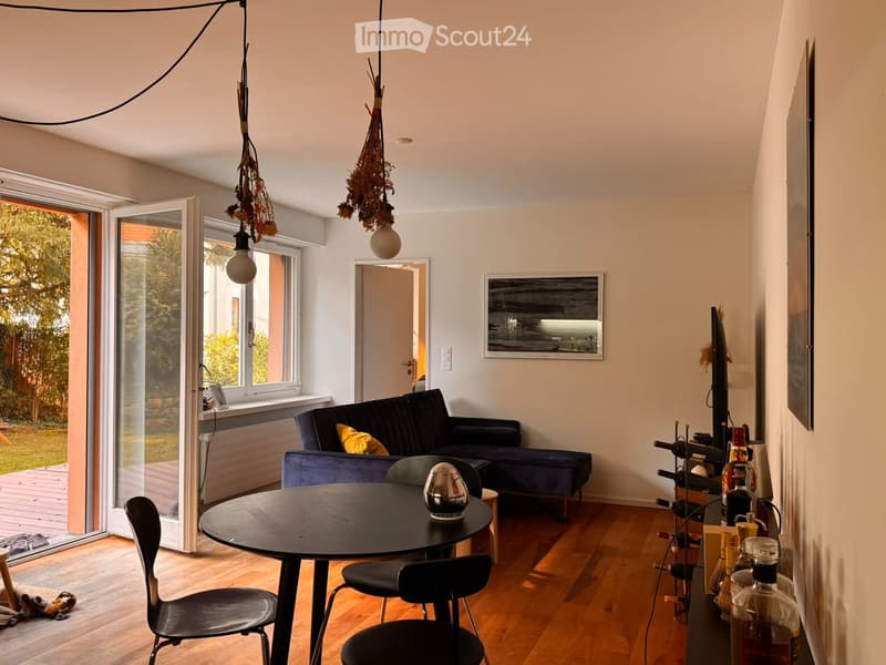 Modern living room with wooden flooring, large windows, a dark blue sofa, a round black table with chairs, and a bar area with bottles and a TV.