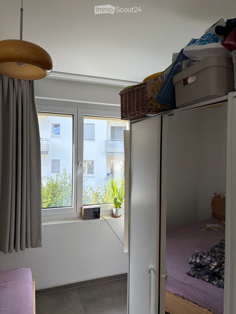 A small bedroom with a bed, a wardrobe, a window with a curtain, a pendant light, a radio on the windowsill, and a potted plant. The wardrobe has baskets on top.