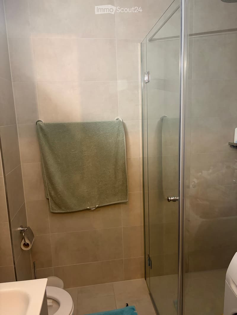 Modern bathroom with beige tiles, a glass-enclosed shower, a toilet, a sink, and a towel hanging on the wall.