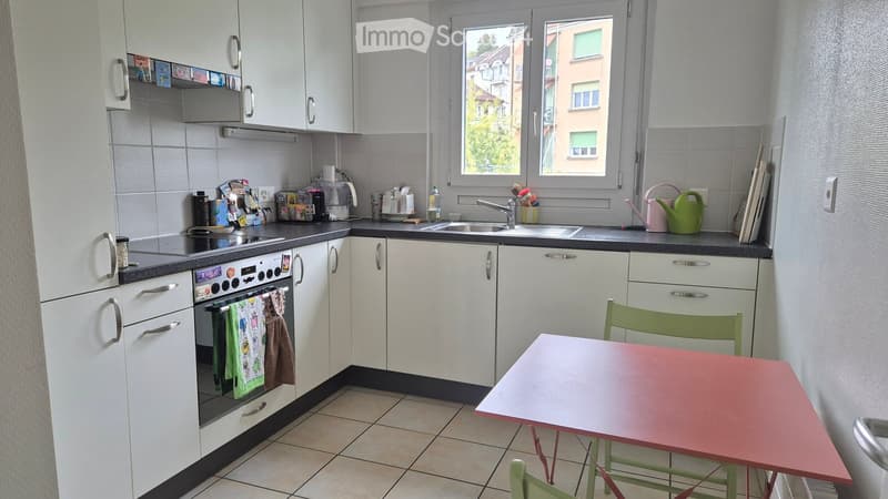 The kitchen features white cabinets, a tiled floor, and a window overlooking the street. It includes an oven, sink, and a small dining area with a table and chairs.