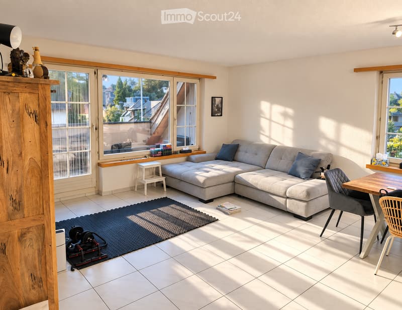 Bright living room with large windows, grey sectional sofa, wooden coffee table, wicker chair, exercise bike, and decorative items on shelves.