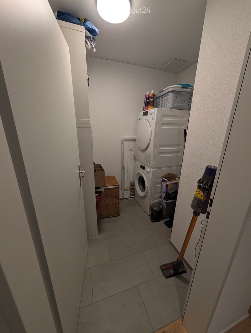 A narrow hallway with tiled flooring, a white washing machine and dryer unit, a vacuum cleaner, and a small woven basket.