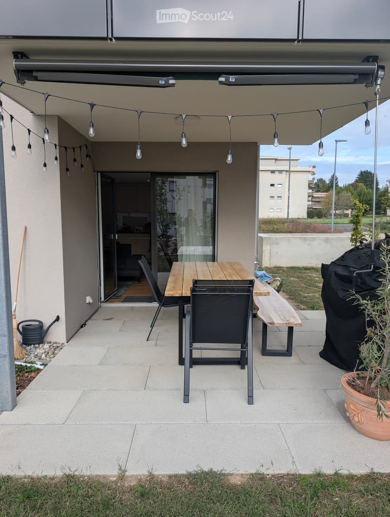 Balcony with a wooden table, two black chairs, a wooden bench, and a potted plant. The area is covered by a retractable awning with string lights.