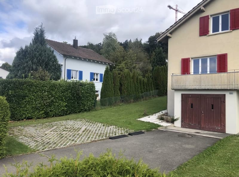 Two-story house with beige and light blue exterior, red shutters, balcony, garage, and well-maintained garden.