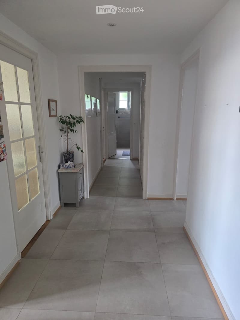 A clean, well-lit hallway with tiled flooring, white walls, and a small side table with a plant. The hallway leads to another room with visible doors and windows.