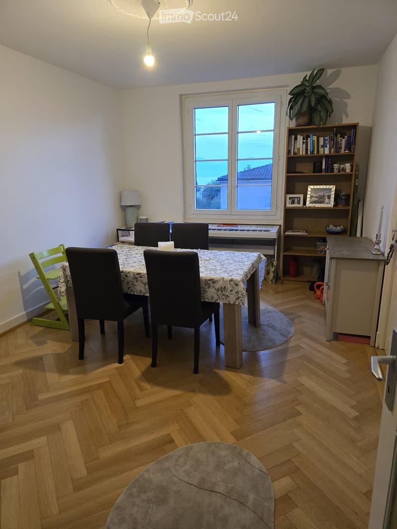 Dining area with wooden herringbone flooring, a round table with four black chairs, a piano, a bookshelf, and a small green chair.