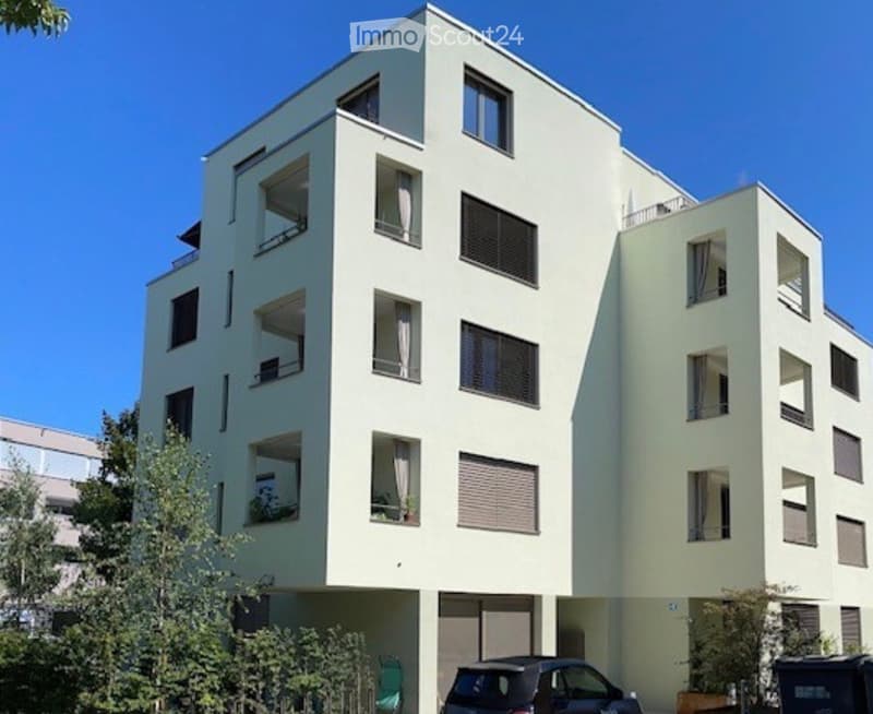 Modern multi-story residential building with light-colored facade, multiple windows, balconies, and a parking area at the base.