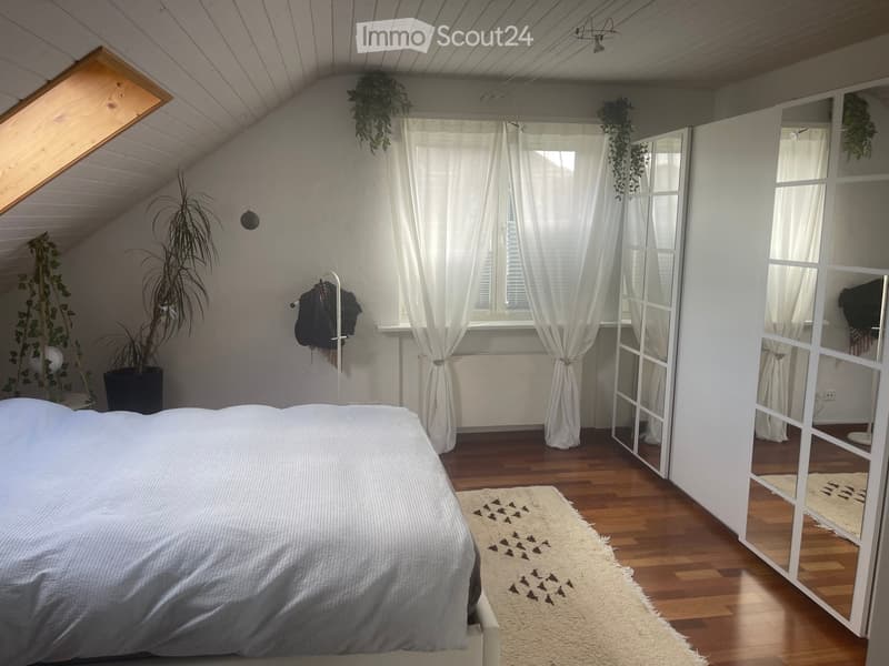 Cozy attic bedroom with wooden floors, large window with white curtains, bed with grey bedding, cream-colored rug with bird pattern, wardrobe with mirrored doors, and several potted plants.