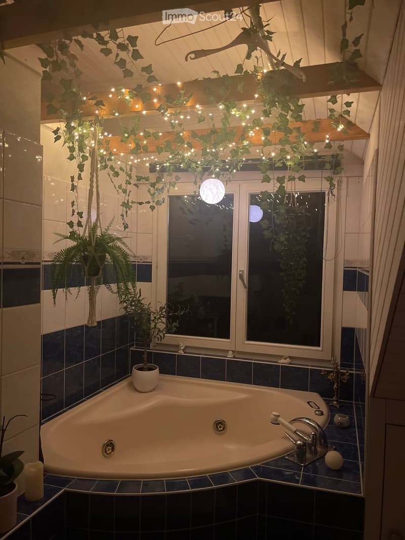 Bathroom with a large bathtub, decorative plants, and string lights hanging from the ceiling. The room features blue tiles and large windows with a view of the night sky.