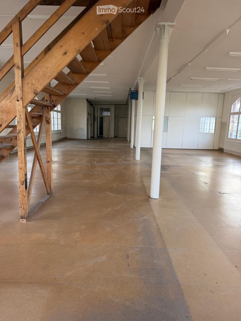 Empty room with wooden floor, white walls, large windows, and a wooden staircase leading to an upper level.