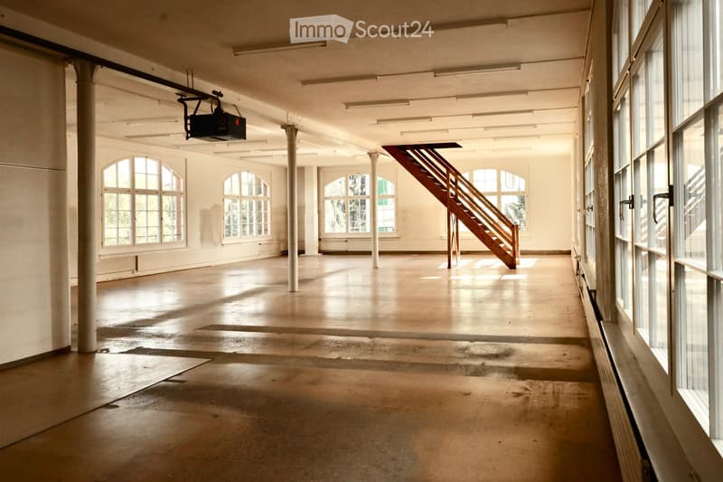 Empty industrial-style room with large windows, concrete floor, exposed metal columns, and a wooden staircase leading to an upper level.