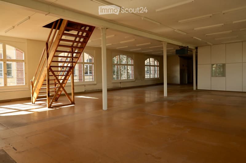 Empty room with wooden floor, large windows, white walls, and a wooden staircase leading to an upper level.