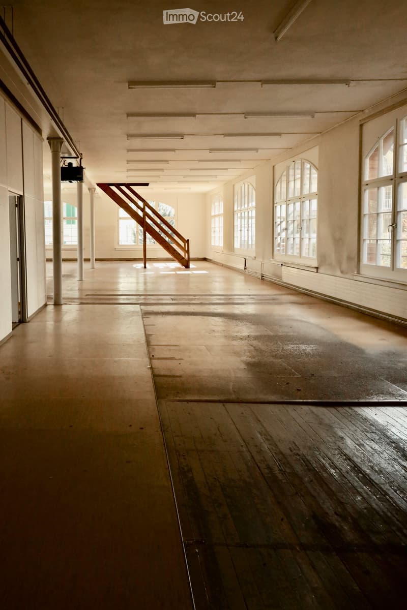 Empty room with wooden floors, large windows, and a staircase leading to another level. The space is well-lit with natural light and has exposed structural elements.