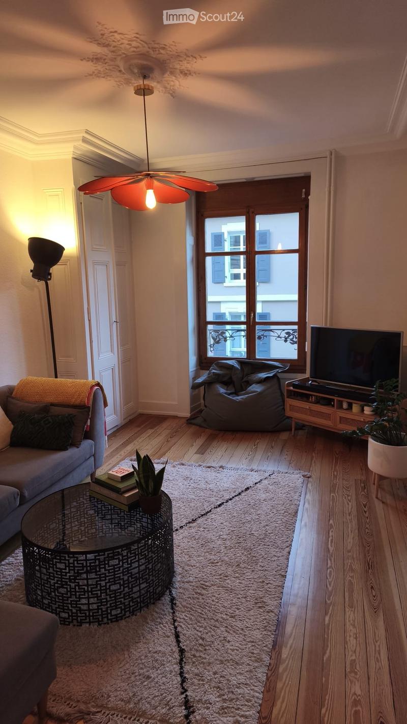 Modern living room with wooden flooring, a large white shag rug, a grey sofa with pillows, a round glass coffee table with books and a plant, a bean bag chair, a TV on a wooden stand, and a hanging pendant light.