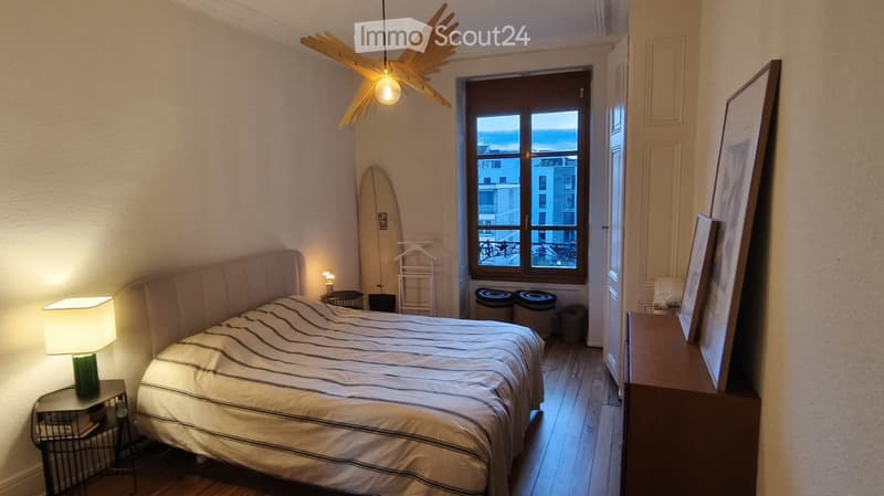 Cozy bedroom with a large bed, nightstand with lamp, picture frame on dresser, surfboard leaning against wall, and a view of the city through the window.