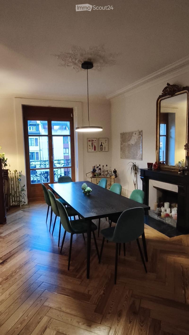 Dining room with a dark wooden dining table, six green chairs, a fireplace with candles, a large mirror, and a small bar cart with bottles.