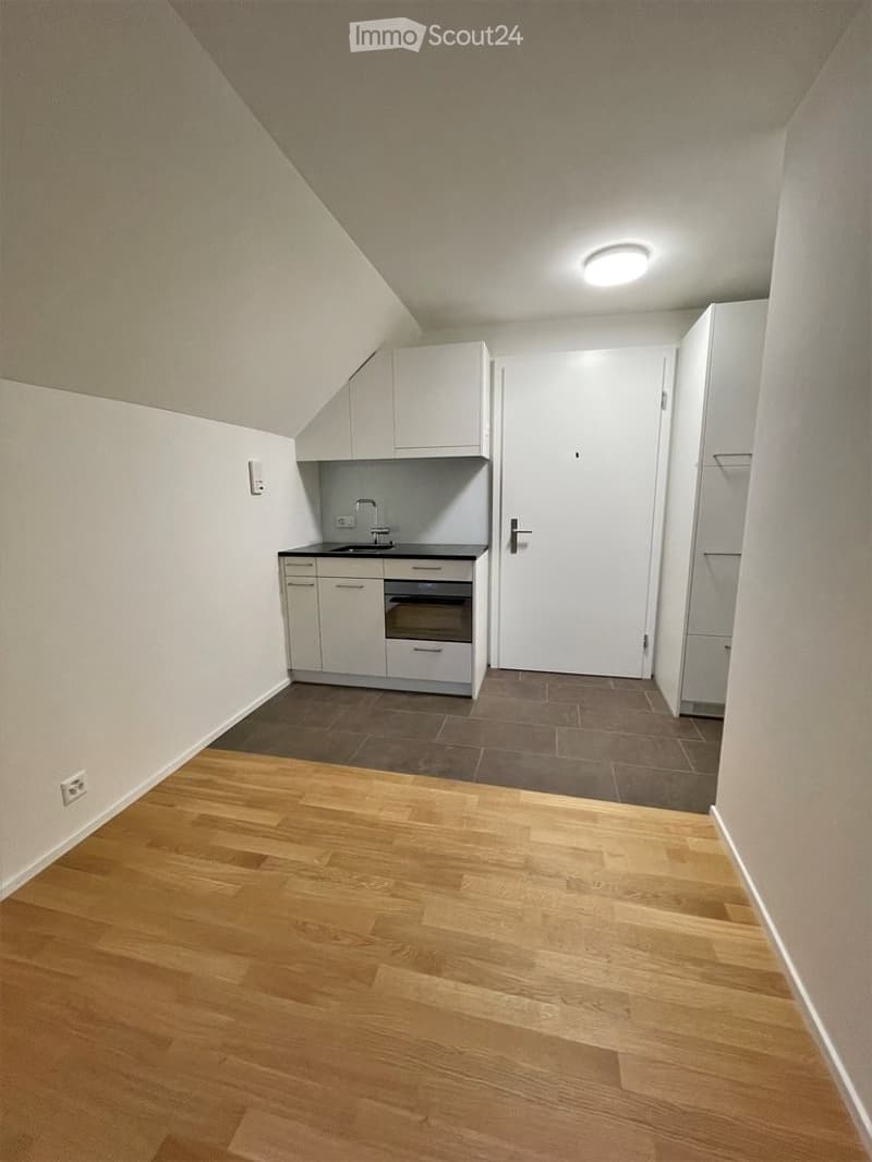 Empty room with wooden flooring, white walls, and a small kitchen area featuring cabinets, sink, and oven.