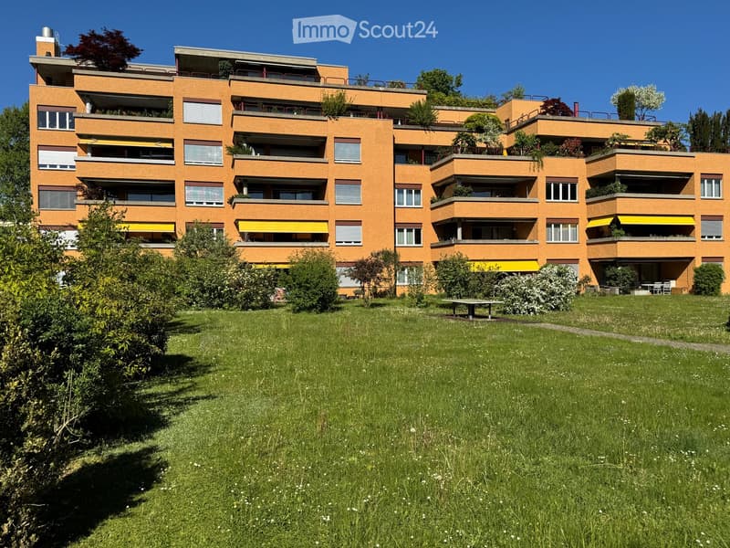 Building. Multi-story orange apartment building with balconies and greenery, surrounded by a grassy lawn and trees.