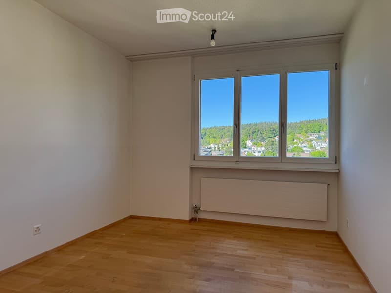 Unfurnished Room. Empty room with wooden flooring, white walls, large window with view of trees and houses, radiator below window, and a single light fixture on the ceiling.
