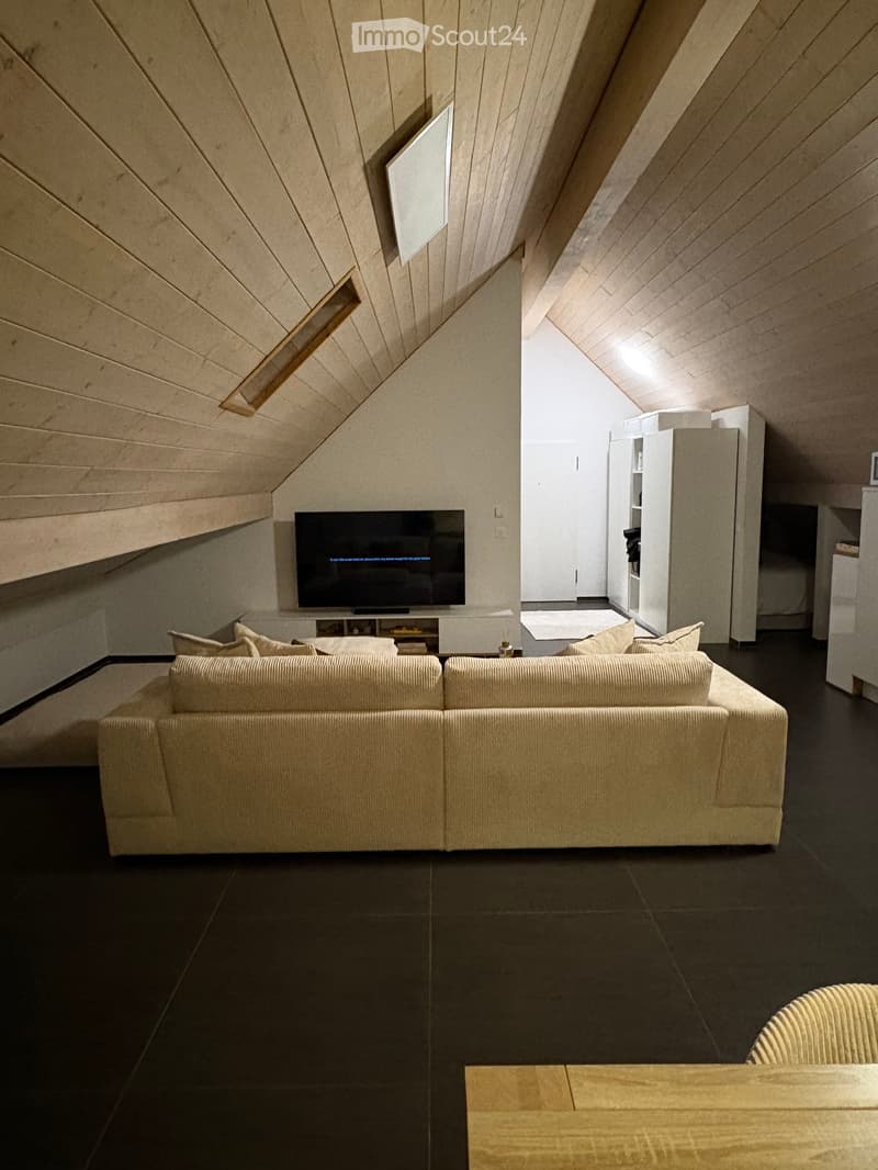 Modern living room with a sectional sofa, coffee table, television, and a wooden chair, featuring dark flooring and a slanted wooden ceiling.