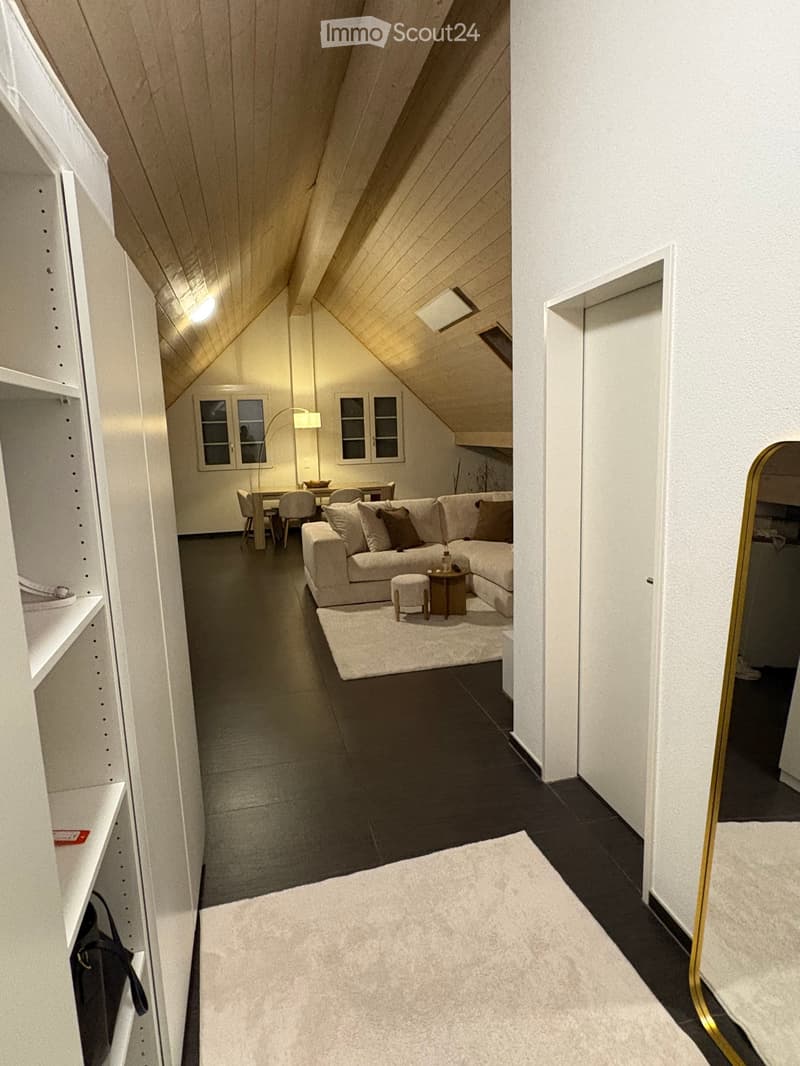 Modern living room with a sectional sofa, coffee table, and dining area in an attic space with wooden slanted ceilings and large windows.