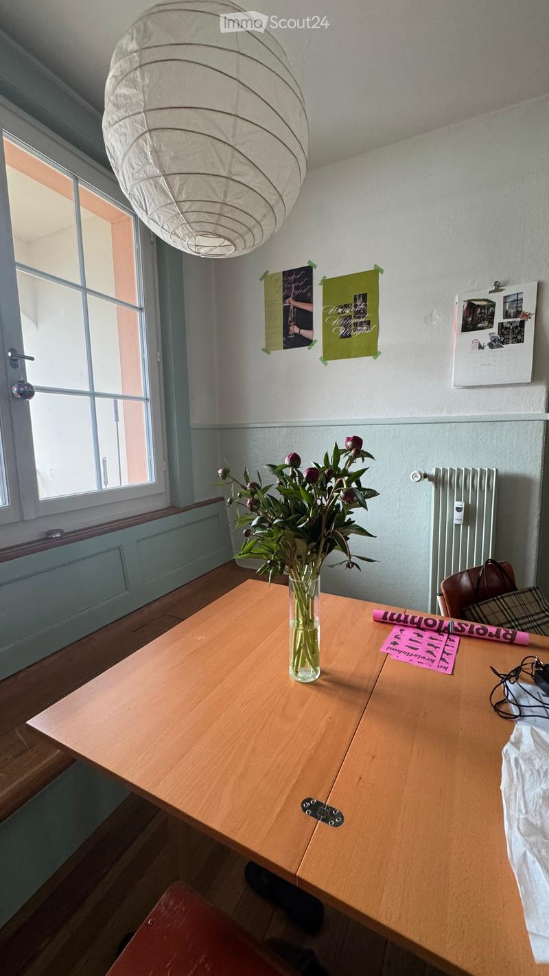 A small, well-lit room with a wooden table, vase of flowers, chair, posters, and a calendar on the wall.
