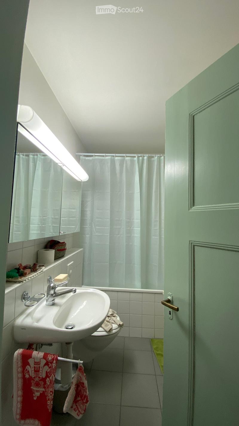 A small, well-lit bathroom with a sink, toilet, mirror, and shower area. The room features white tiles, a white shower curtain, and a towel hanging on a rack.