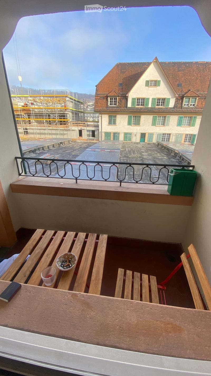 Balcony with wooden bench, cup, and book, overlooking a courtyard with a building and construction site.