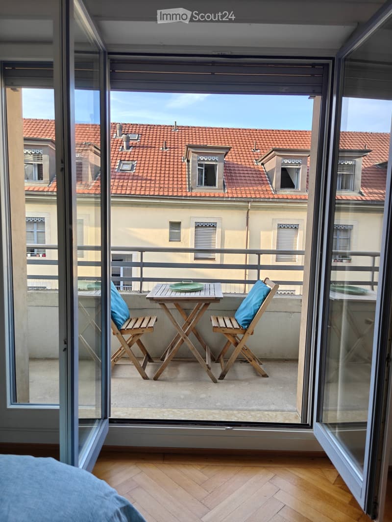 Balcony with a small wooden table and two chairs, overlooking a cityscape with red-tiled roofs.