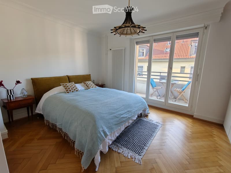 Cozy bedroom with wooden flooring, a large bed with a blue duvet and patterned pillows, a nightstand with a lamp and vase, and a view of a balcony with outdoor chairs.
