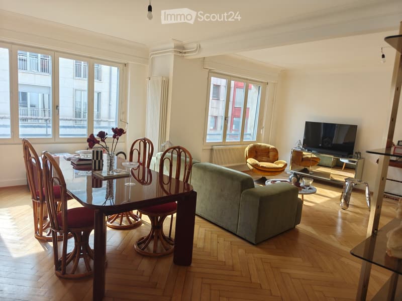 Bright living room with wooden flooring, large windows, a green sofa, a wooden dining table with chairs, a television, and decorative items.