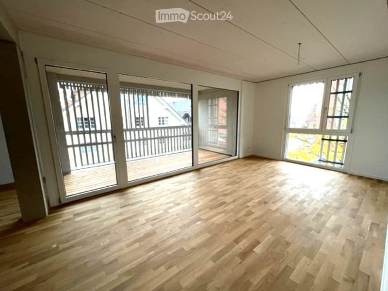 Empty room with wooden flooring, large windows, and sliding glass doors leading to a balcony.