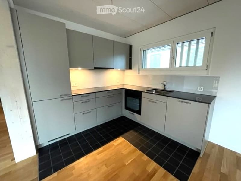 Modern kitchen with grey cabinets, black tiled floor, and wooden accents. Features a sink, oven, and built-in lighting.