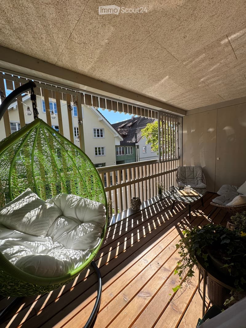 Balcony with wooden decking, a wicker hanging chair, a small table, a chair, and potted plants.
