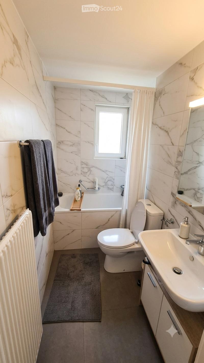 Modern bathroom with marble tiles, a bathtub, toilet, sink, mirror, and a towel on a rack. The room features a window with a curtain and a radiator on the wall.