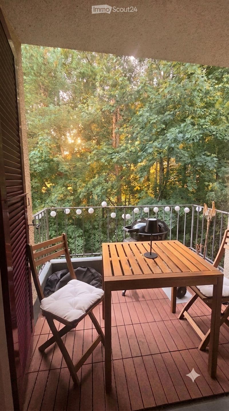 Balcony with wooden table, chairs, and a grill, surrounded by greenery.