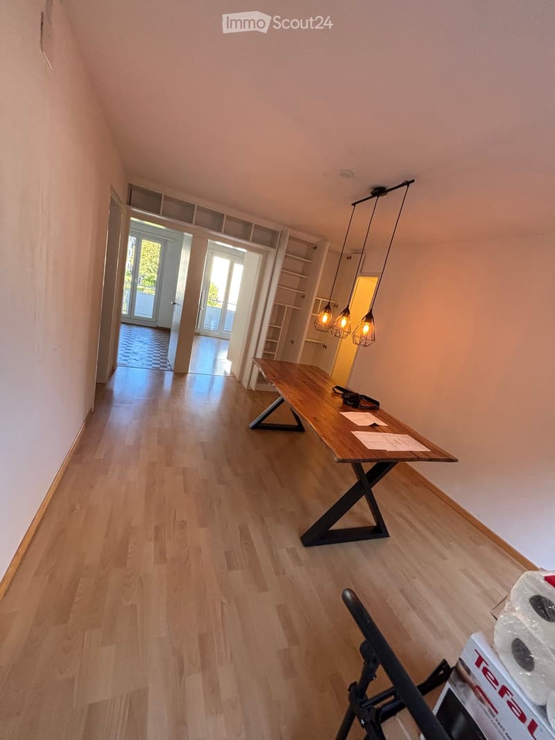 A modern hallway with light wooden flooring, a sleek wooden table with a geometric base, and a hanging pendant light fixture. The hallway leads to an open area with large glass doors and a view of greenery outside.