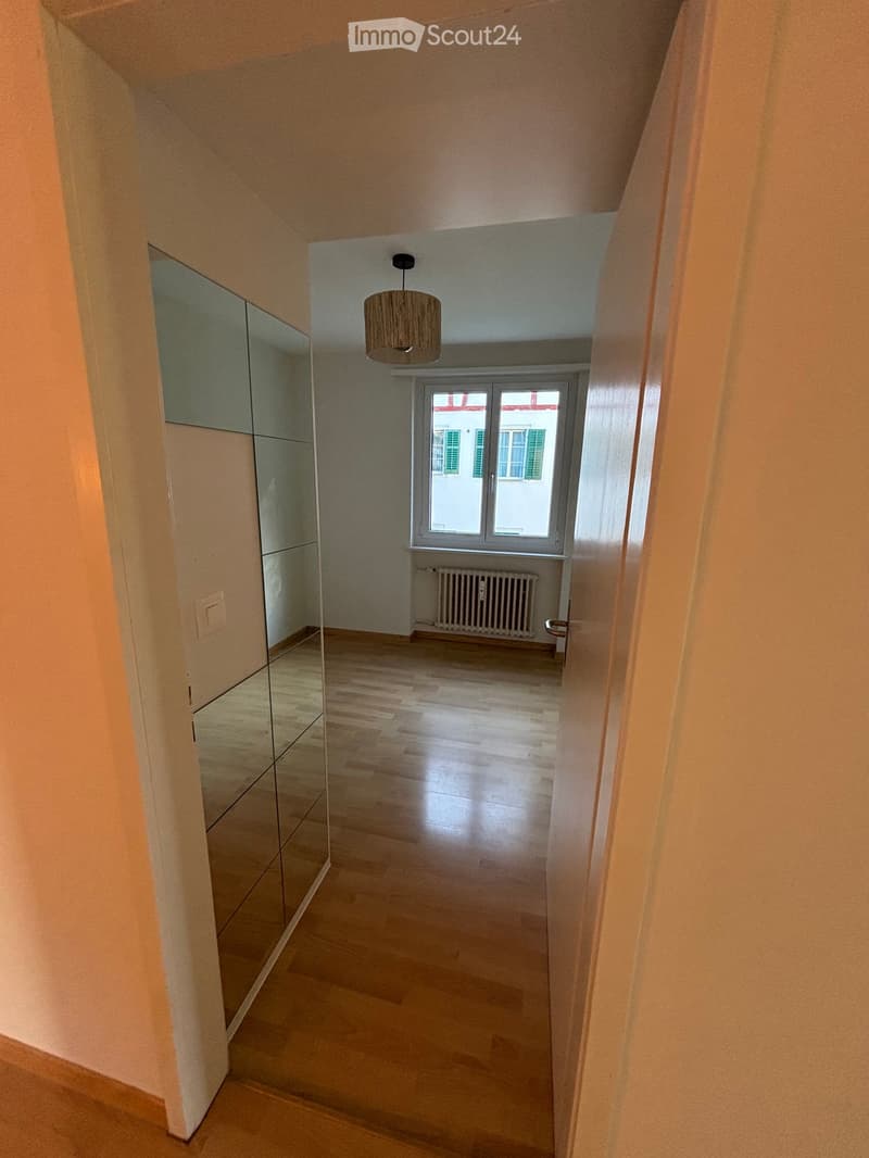 Empty hallway with wooden flooring, a large mirror on one wall, a hanging light fixture, and a window at the end showing a view of another building.