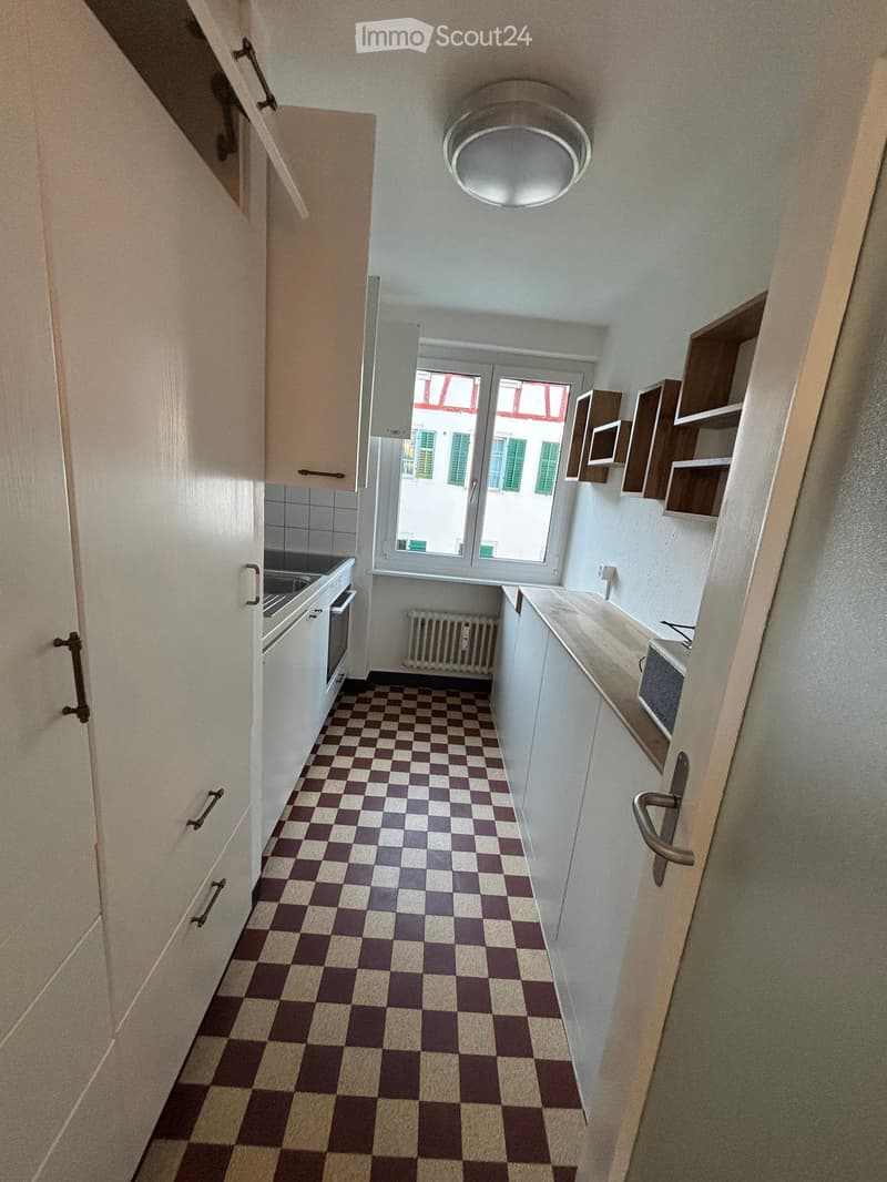 Compact kitchen with checkered floor, white cabinets, stove, oven, sink, radiator, and air conditioner. Features a window with a view of a building with green shutters.