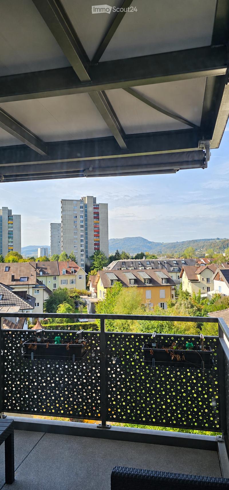 Balcony with modern railing, potted plants, and outdoor furniture overlooking a cityscape with residential buildings and distant mountains.