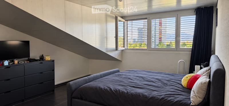 Modern bedroom with a large window offering a view, a bed with pillows, a nightstand, and a television on a dresser.