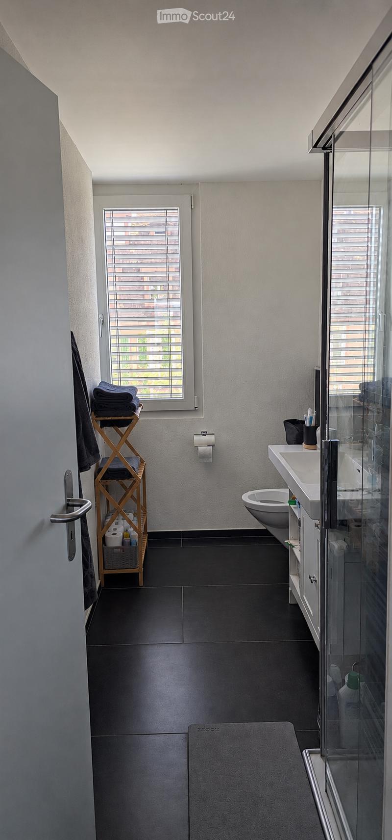 Modern bathroom with dark tiled floor, white sink, toilet, towel rack, and a glass shower enclosure. The room features a window with blinds and a small wooden shelf holding towels and toiletries.