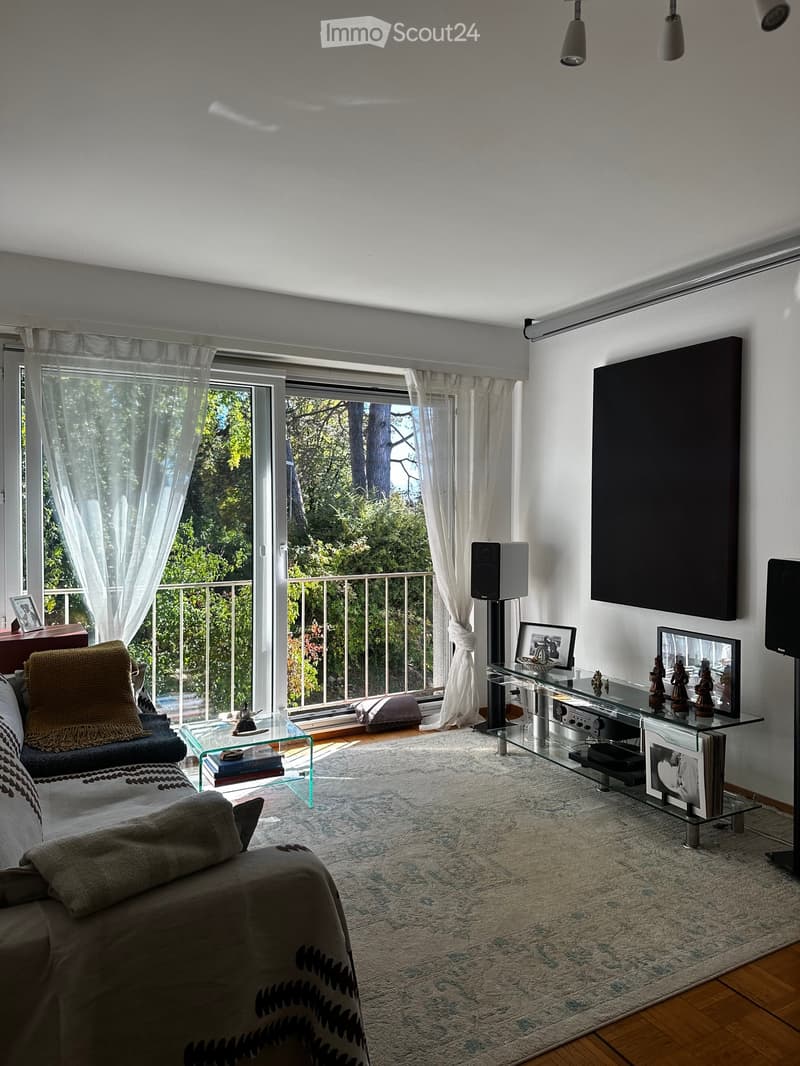 Modern living room with large windows and balcony access, featuring a sofa, coffee table, glass side table, speaker system, and decorative items.