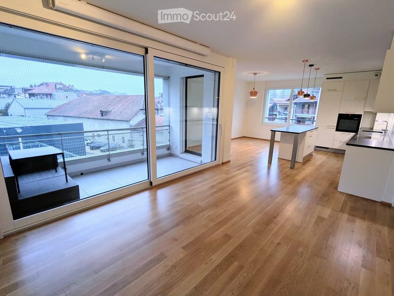 Modern, open-plan living and kitchen area with light wooden flooring, large sliding glass doors leading to a balcony, and contemporary kitchen appliances.