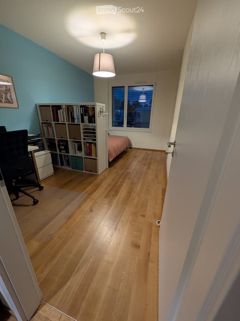 A well-lit bedroom with wooden flooring, a bed with pink bedding, a bookshelf filled with books, a desk chair, and a hanging lamp.