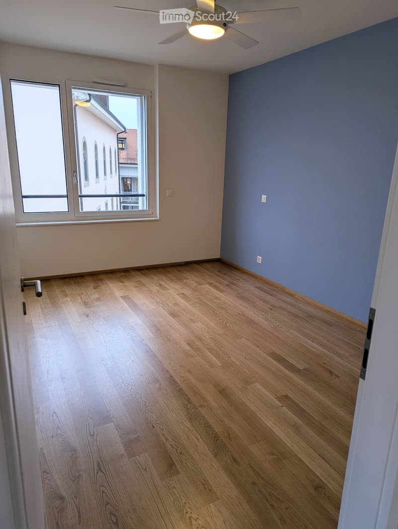 Empty room with light wooden flooring, white walls, and a large window with a view of a building outside. The ceiling features a modern light fixture.